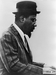 Thelonious-Monk-gettyimages-51957245watermark Man in checked jacket wearing a porkpie hat and glasses playing piano.