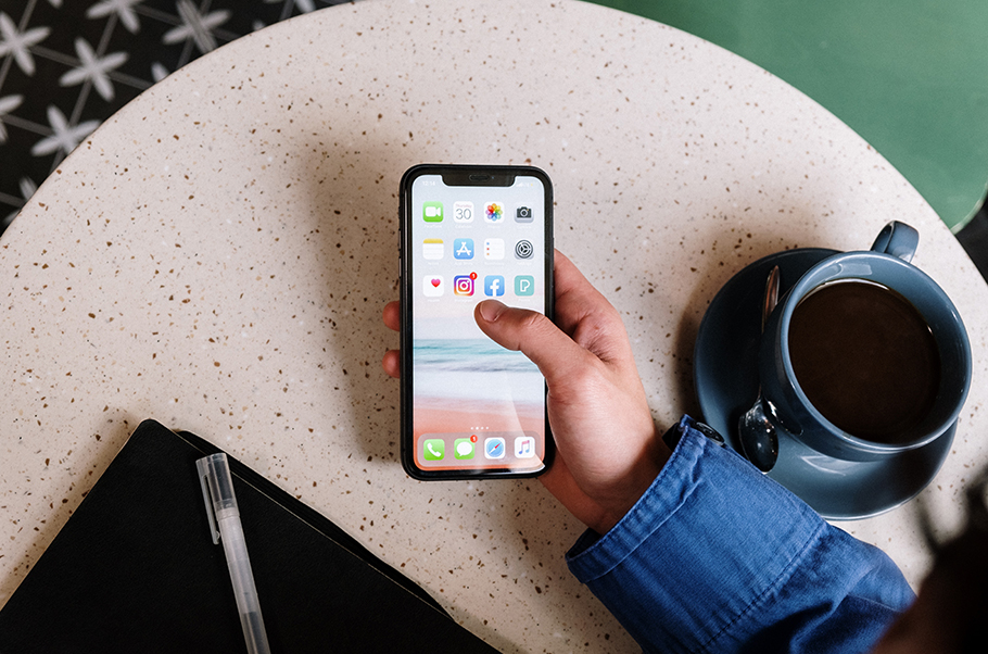 Someone's hand holding an iphone at a table where there is a cup of coffee and a notepad with pen.