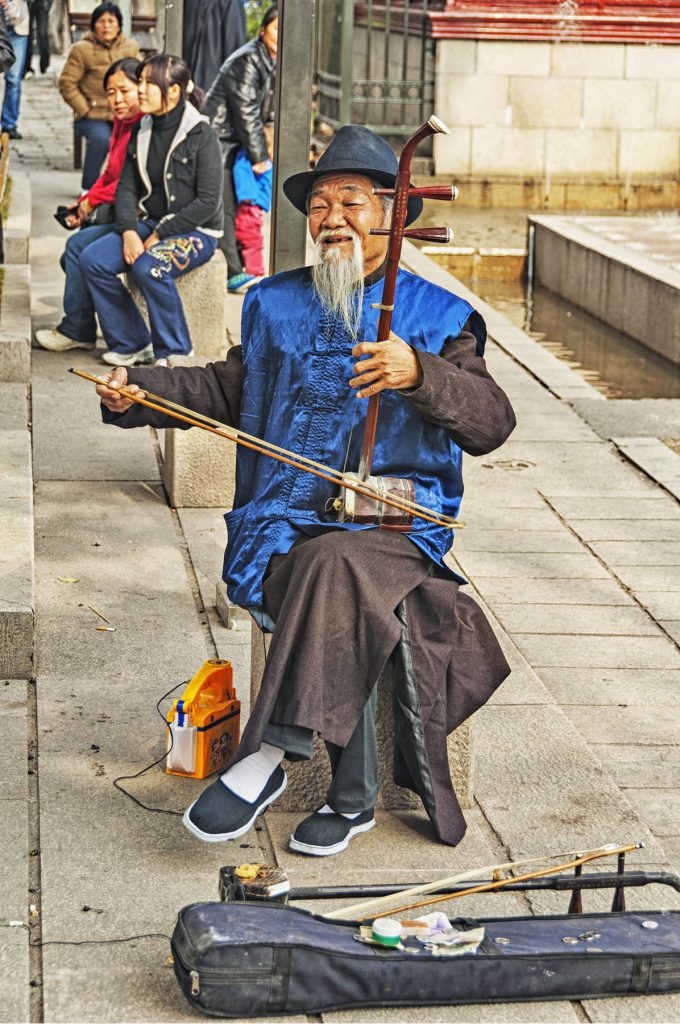 Erhu-reduced Elderly man with long beard in traditional dress sitting in a plaza playing a stringed instrument with a bow.